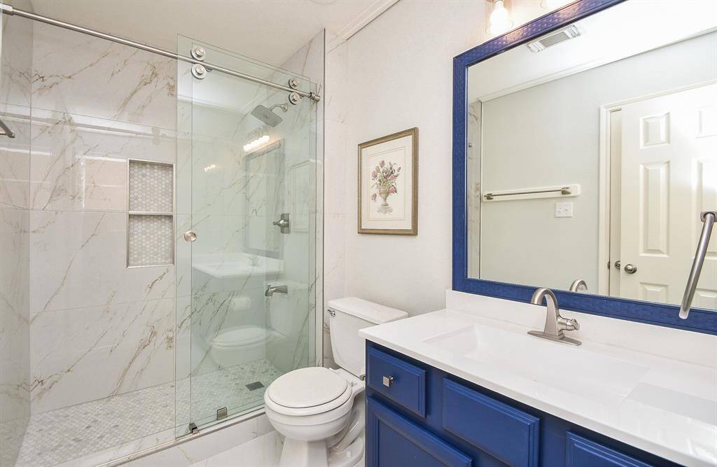 This bathroom features a sleek, modern design with a glass-enclosed walk-in shower, marble-style tiles, and built-in shelving. The vanity includes a bold blue cabinet with a white countertop and a large mirror, adding a pop of color and elegance.