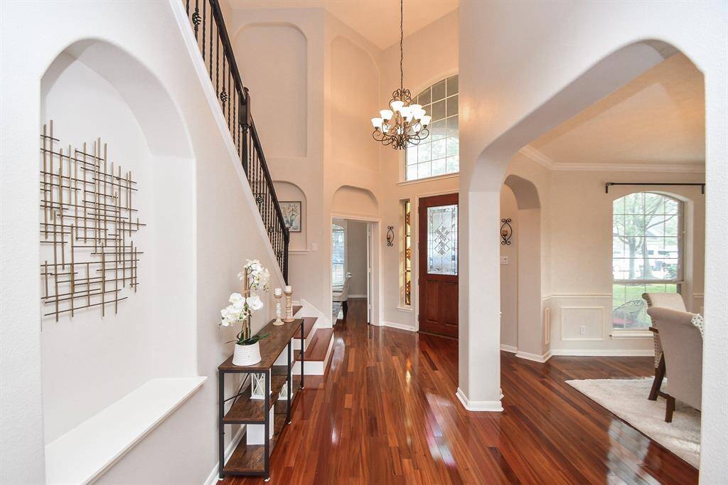 This elegant home entryway features high ceilings, a grand chandelier, and warm hardwood floors. Arched doorways lead to a bright, inviting living space with large windows. A staircase with wrought iron railings adds a touch of sophistication.