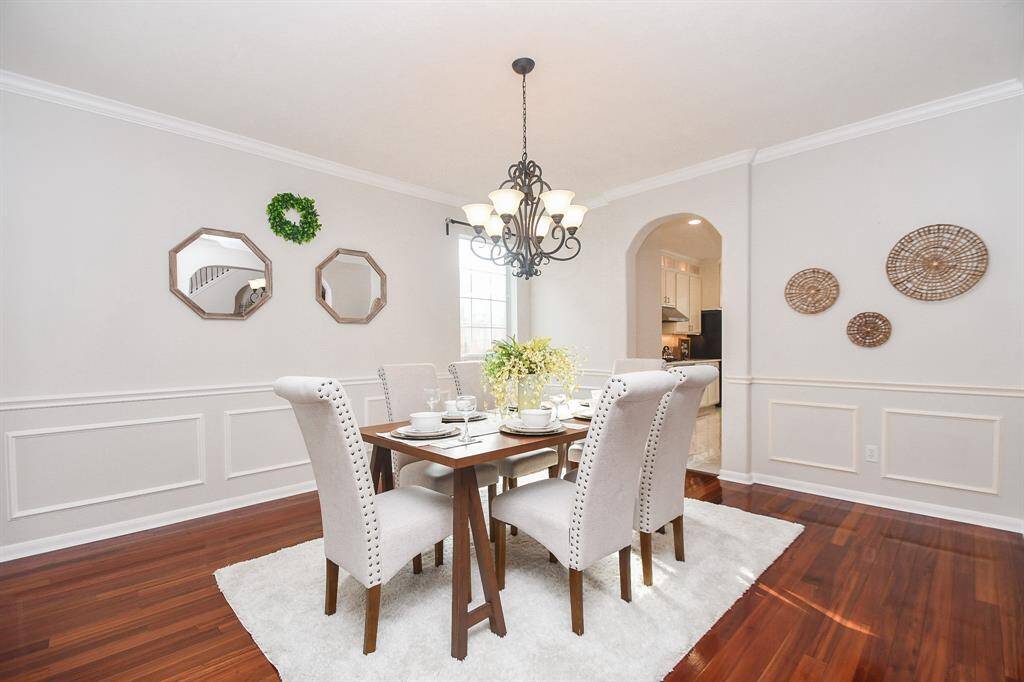 This elegant dining room features rich hardwood floors and wainscoting, with a stylish chandelier above the table. The space is brightened by a large window and decorated with wall accents, creating a warm and inviting atmosphere.
