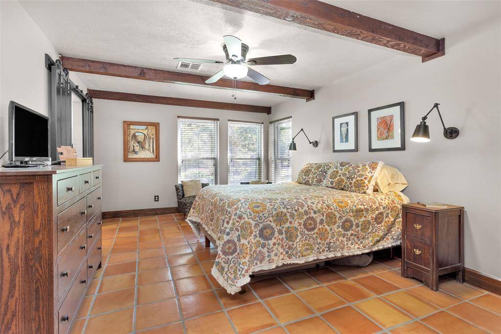 The primary bedroom continues the home's rustic charm, showcasing wood beams, swing-arm reading lights inspired by bank teller designs, and sliding panel doors leading to the primary bathroom. The corner windows fill the room with natural light.