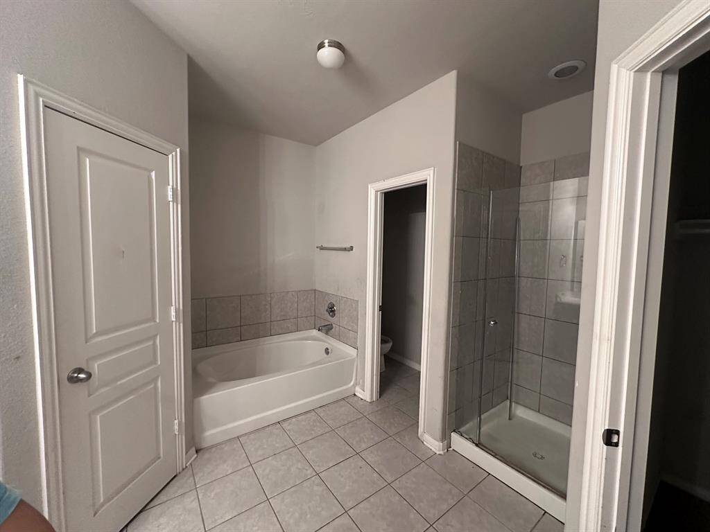 This bathroom features a clean, modern design with a bathtub, separate shower, and tiled flooring. There are white walls, a ceiling light, and a door leading to a toilet area.
