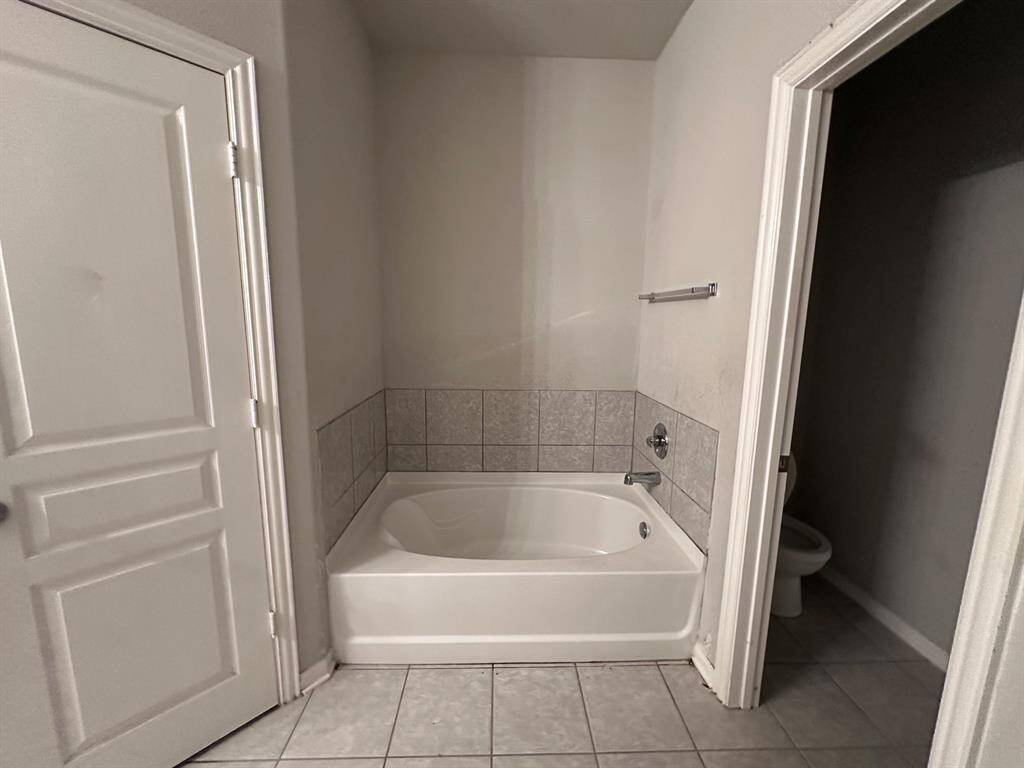 The photo shows a bathroom with a white bathtub, tiled walls, and a tiled floor. There's an adjacent door leading to a toilet area, and another door on the left side of the image.