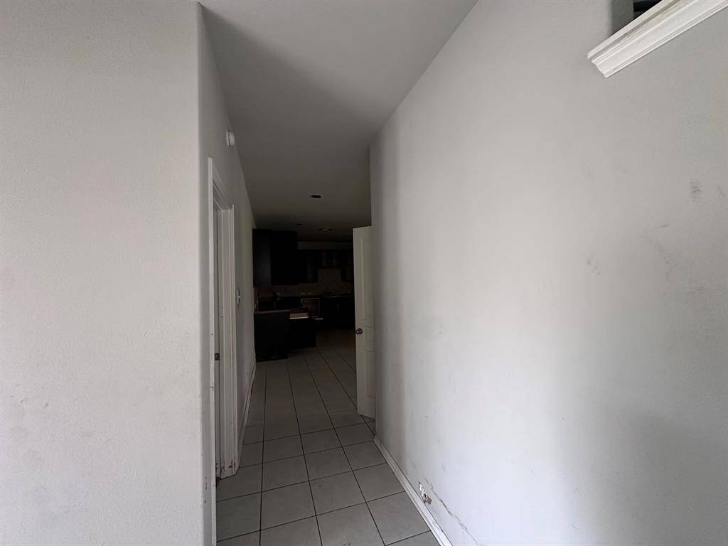 This photo shows a simple hallway with light-colored walls and tiled flooring, leading to a kitchen area. The space appears narrow and has a door on the left.