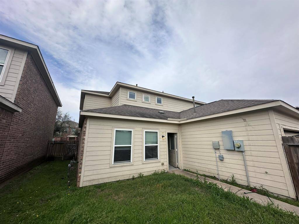 This is a photo of the backyard of a two-story house with light-colored siding. It features a small grassy area, fenced on one side, with visible utility boxes on the exterior wall. The house has a simple roof design and several windows, providing natural light inside.