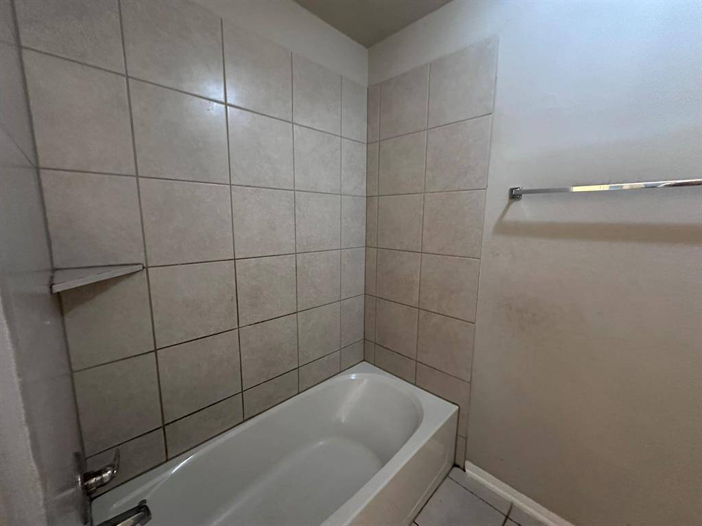 This bathroom features a tiled bathtub and shower area with neutral tones, providing a clean and simple look. There's a wall-mounted towel bar and a small built-in corner shelf for convenience.