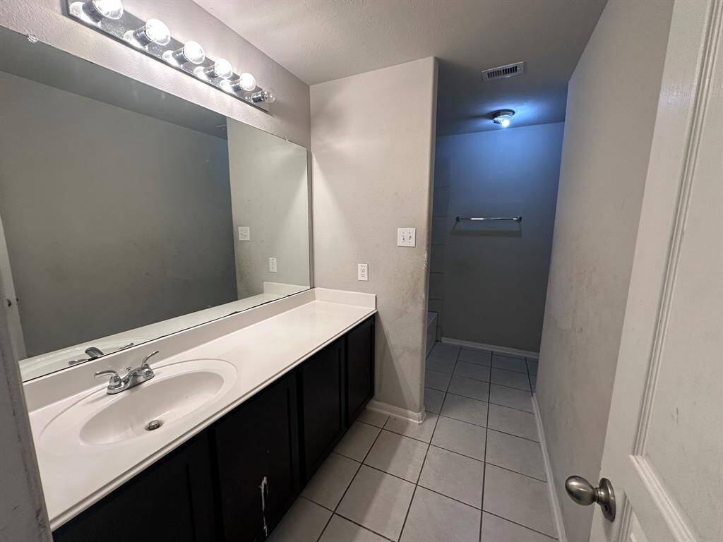 This bathroom features a large mirror with bright lighting, a single-sink vanity with storage, tiled flooring, and a separate shower area.