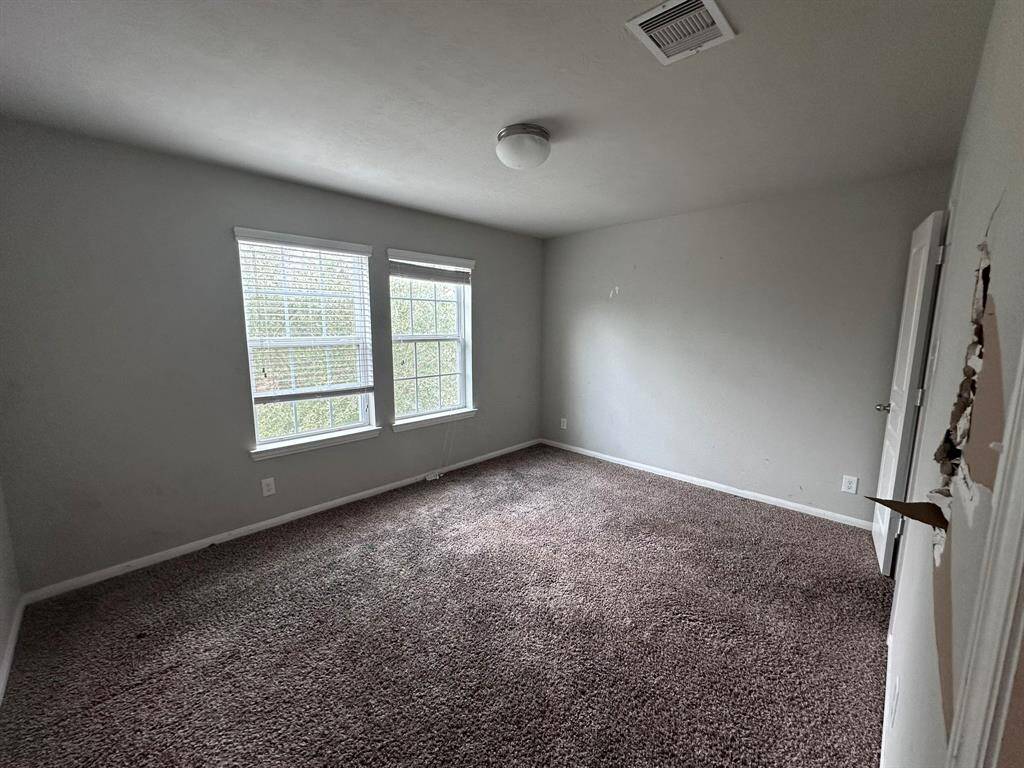 This room features neutral walls, carpeted flooring, and ample natural light from two windows. It has a simple ceiling light fixture and a visible air vent. There is some wall damage near the door.