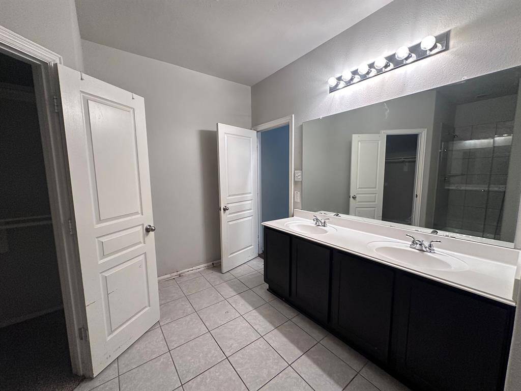 This photo shows a bright bathroom featuring a double vanity with dark cabinets and a large mirror. It has tiled flooring, modern lighting, and two open doors leading to a closet and shower area.
