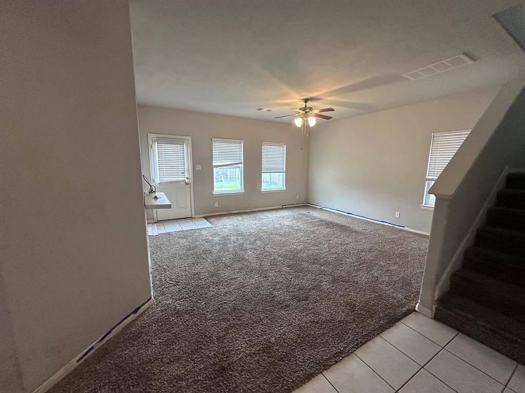 This photo shows a spacious living room with carpet flooring and a ceiling fan. It features multiple windows for natural light and an adjacent tiled entryway. There's a staircase leading to the upper level.