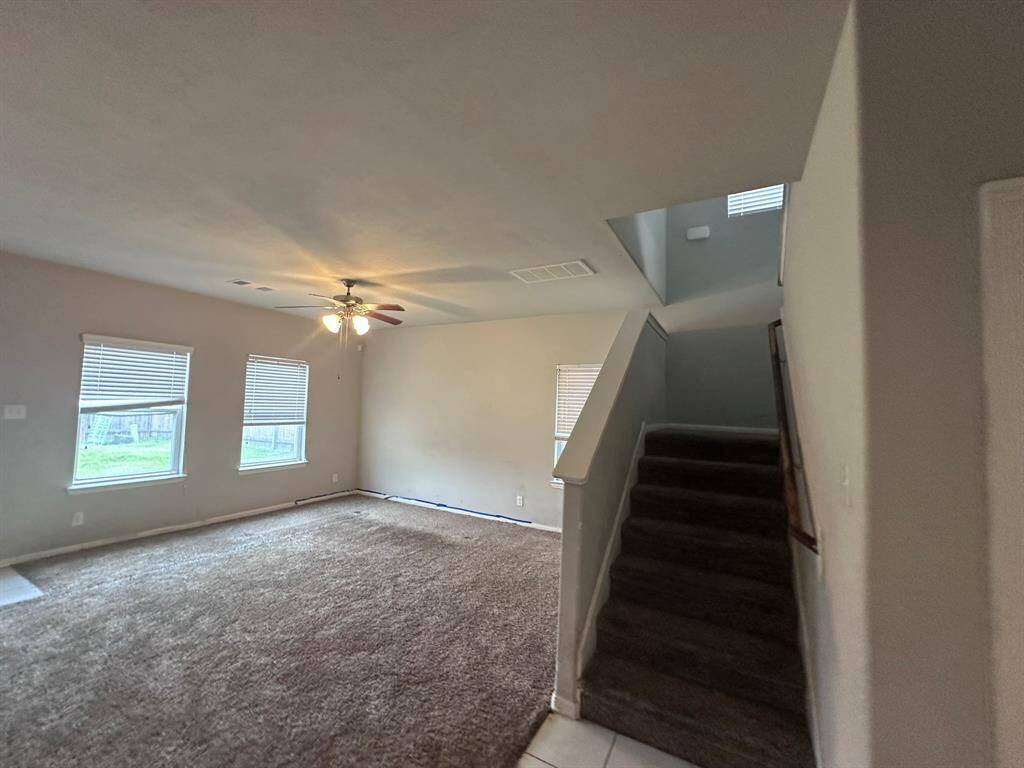 Spacious living area with carpeted flooring, large windows for natural light, a ceiling fan, and a stairway leading to the upper floor.