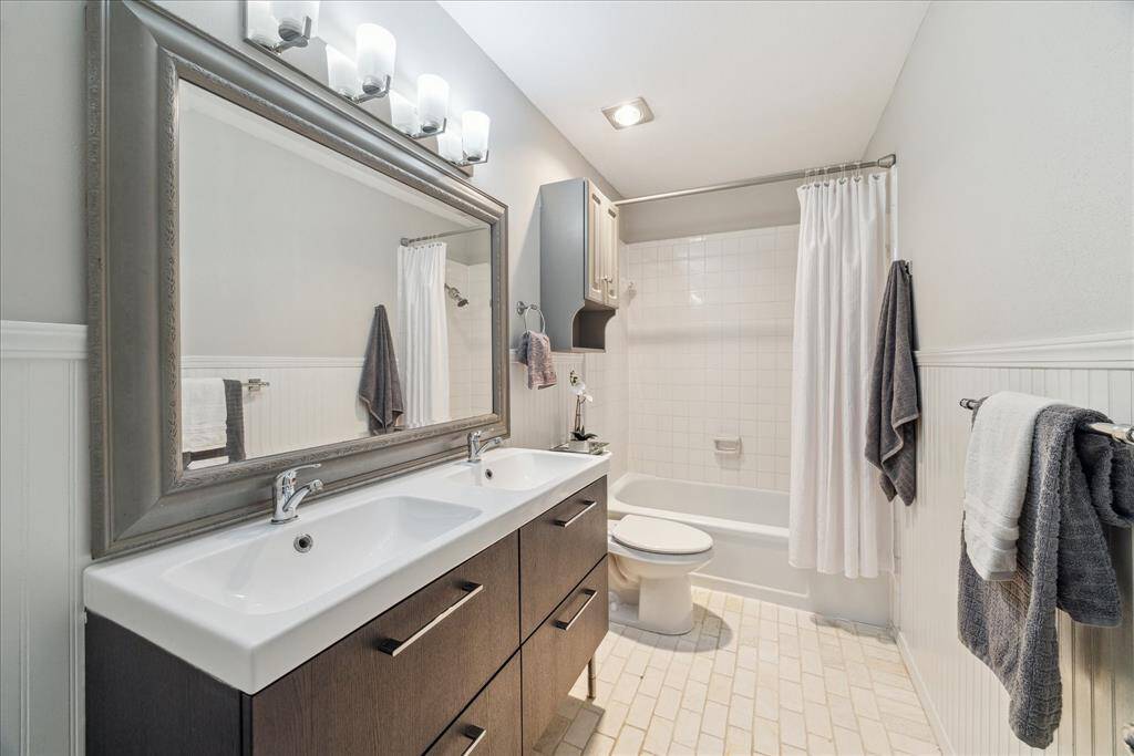 Primary bathroom has been updated with stone tile floors, oversized mirror, modern lighting, heat lamps, wainscoting, and furniture-style double-sinked vanity with storage drawers.
