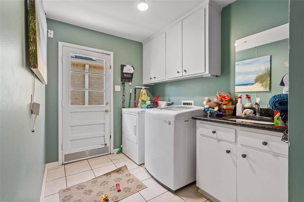 This functional laundry room offers ample storage and workspace with upper and lower white cabinetry, a built-in sink, and granite countertops. The washer and dryer are conveniently positioned, while the door provides easy access to the backyard. The space is accented by green walls, tiled flooring, and a practical layout designed for efficiency.