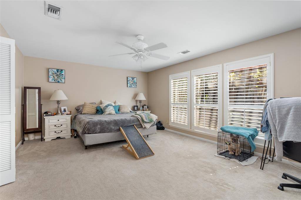 This spacious primary bedroom offers a serene retreat with soft neutral tones, plush carpeting, and abundant natural light streaming through large windows adorned with plantation shutters. The generous layout provides ample space for relaxation, while the ceiling fan ensures year-round comfort.