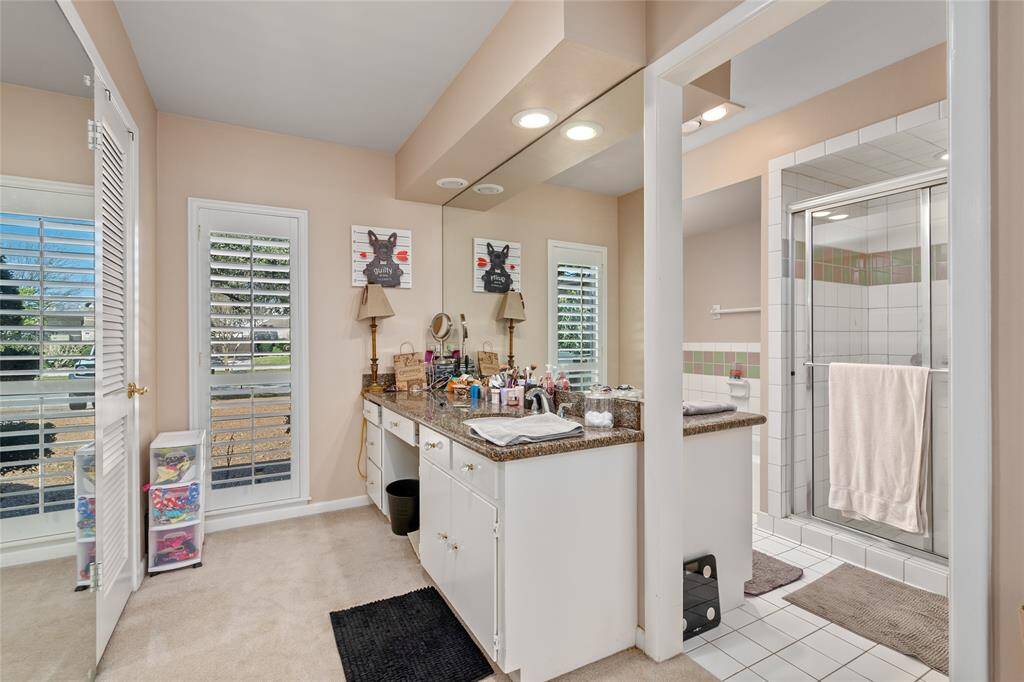 The primary bathroom offers a spacious vanity with ample counter space and storage. A large mirror and built-in lighting enhance the functionality of the space. The ensuite includes a walk-in shower with a glass enclosure, accented by decorative tile. Natural light streams in through the windows with plantation shutters, creating a bright and inviting atmosphere.