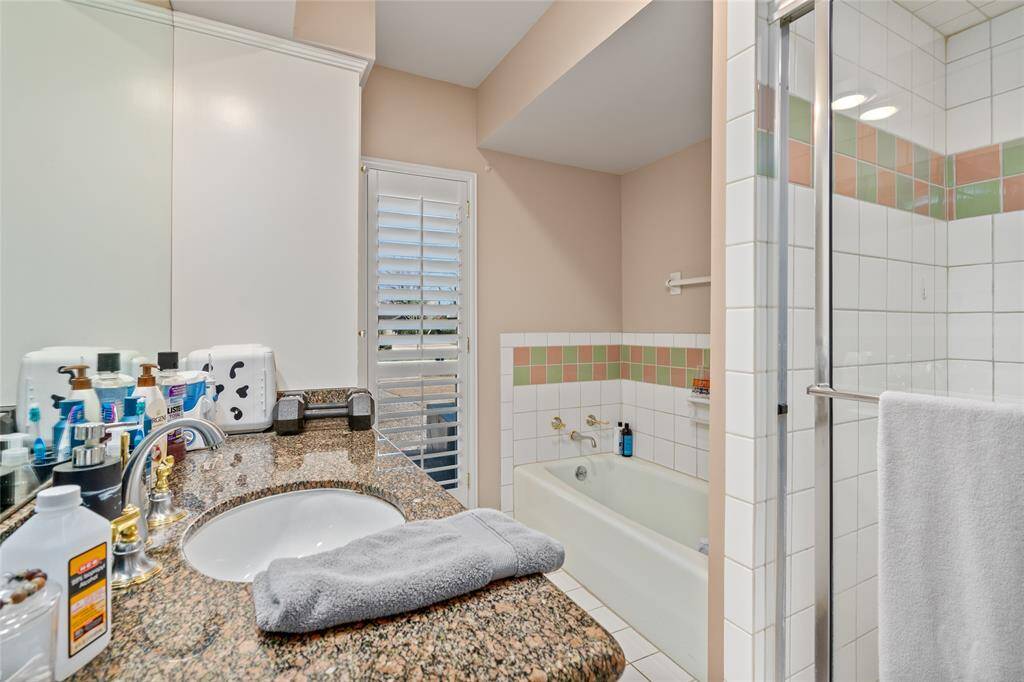 This bathroom features a spacious layout with a combination of a built-in bathtub and a separate glass-enclosed shower. The walls are adorned with neutral tones, complemented by tiled accents. A large vanity area offers ample counter space, and a window with plantation shutters allows for natural light and ventilation.