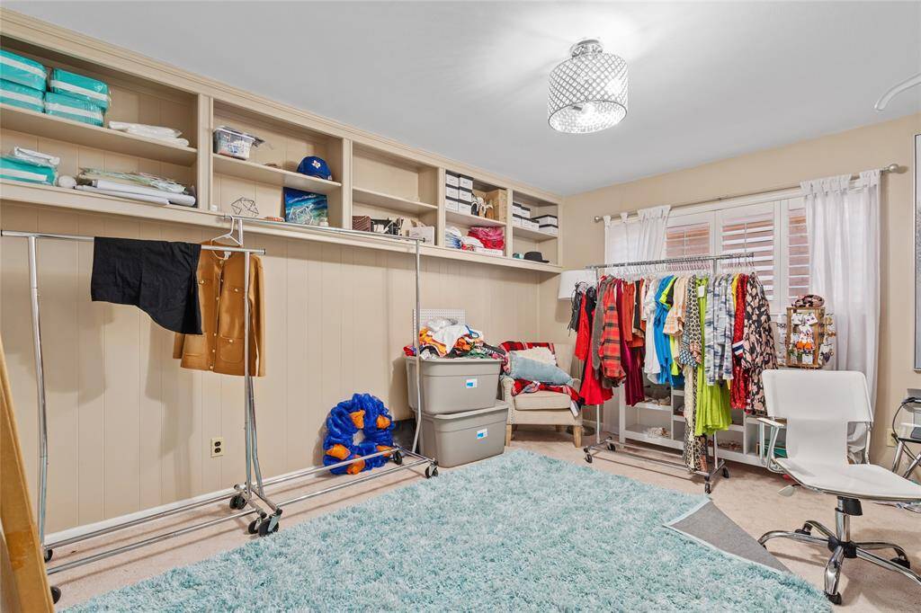 This room features a spacious layout with neutral-toned walls and a light-colored carpet. A large window with plantation shutters allows for ample natural light. Built-in open shelving along one wall provides additional storage, and a ceiling-mounted light fixture brightens the space.