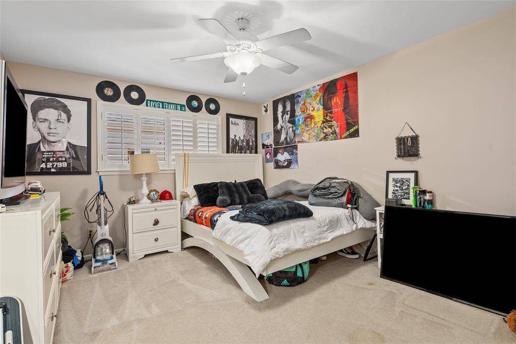 This bedroom features a neutral color scheme with soft carpeting and light-colored walls. A large window with plantation shutters allows natural light to brighten the space. The ceiling is equipped with a fan and light fixture for added comfort. The room offers ample wall space for personalization and decoration.