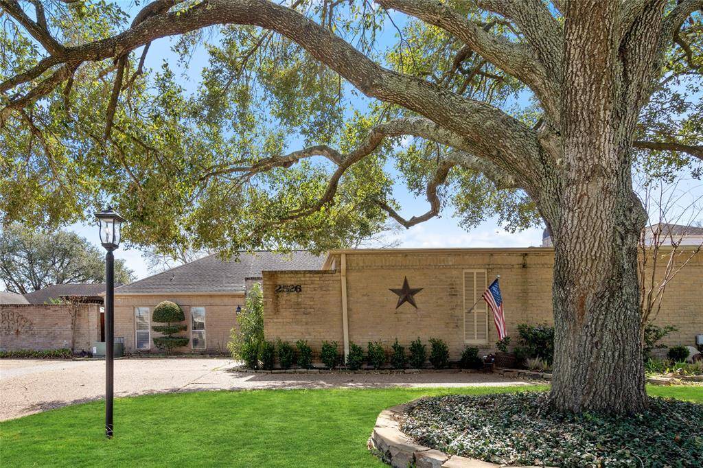 This front yard view highlights a mature tree providing shade and character to the property. A well-maintained lawn and landscaped garden beds enhance the curb appeal. The home’s brick exterior and neatly trimmed hedges create a classic and inviting entrance. A paved driveway leads to the garage and entryway.