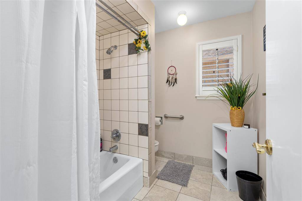 This secondary bathroom includes a tiled shower and bathtub combination, accented with decorative tile inlays. A window with plantation shutters allows for natural light while maintaining privacy. The neutral-toned walls and flooring create a warm and inviting atmosphere. A built-in grab bar adds accessibility, and the layout provides ample space for storage and functionality.