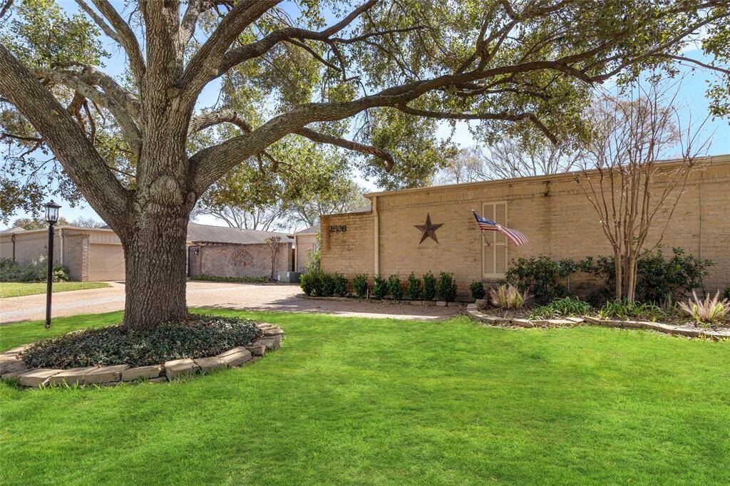This front yard features a lush green lawn and a striking mature tree that provides ample shade. A well-defined landscaping bed surrounds the base of the tree, complementing the neatly arranged shrubbery along the home's brick exterior. The spacious driveway offers easy access to the garage and entryway.