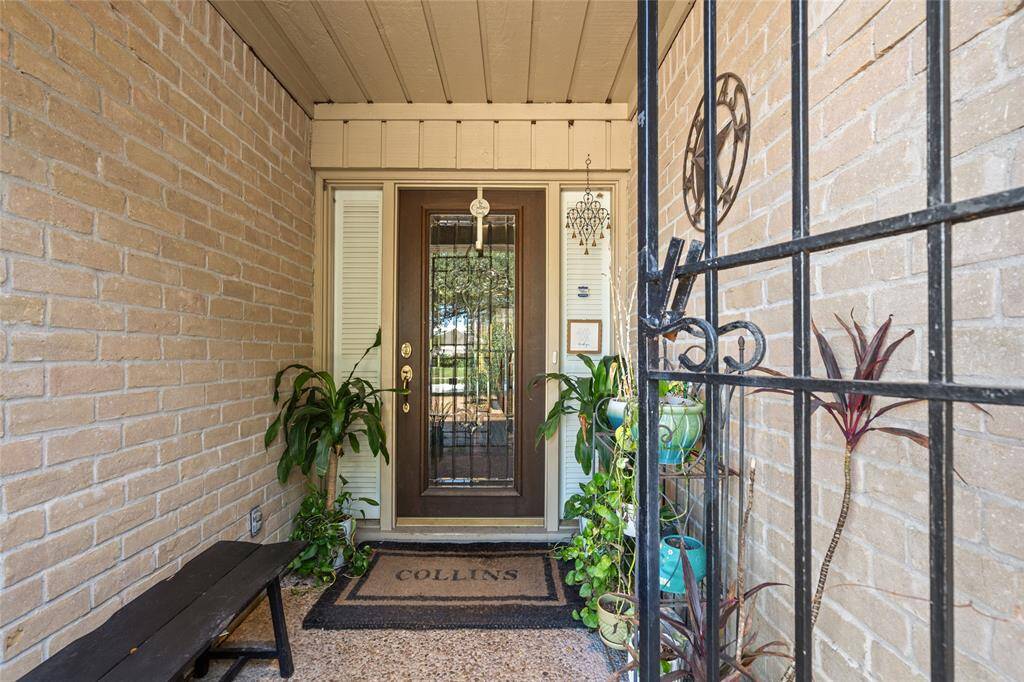 The welcoming front entryway features a charming brick exterior and a decorative iron gate for added security. A glass-paneled door allows natural light to filter in, creating a warm and inviting atmosphere. Lush greenery and potted plants enhance the cozy ambiance, while the covered porch provides a shaded retreat.