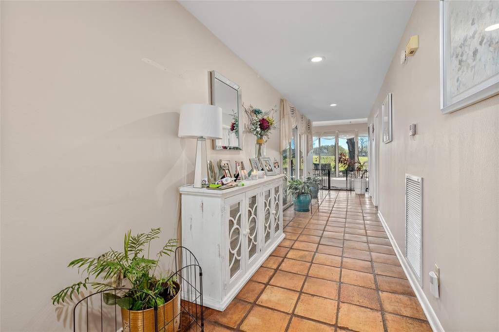 A long, inviting hallway with warm-toned tile flooring leads into the home, offering a seamless flow toward the main living spaces. Neutral walls and recessed lighting create a bright and airy feel, while large windows at the far end frame stunning views of the outdoors.