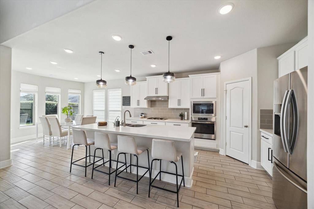 This modern kitchen features a spacious open layout with a large island, bar seating for 5, and pendant lighting. It has stainless steel appliances, white cabinetry, and a cozy dining area with ample natural light from multiple windows to view the pool.