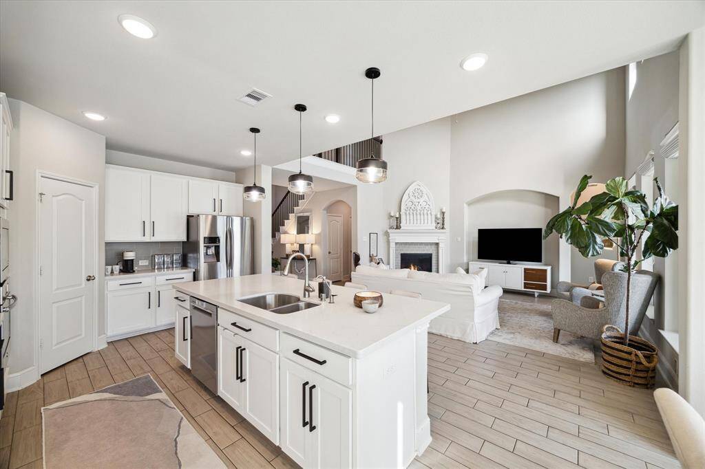This photo showcases a spacious, open-concept kitchen and living area. The kitchen features modern white cabinetry, stainless steel appliances, and a large island with a sink. The living room includes a cozy fireplace, high ceilings, and abundant natural light, creating a welcoming and airy atmosphere.