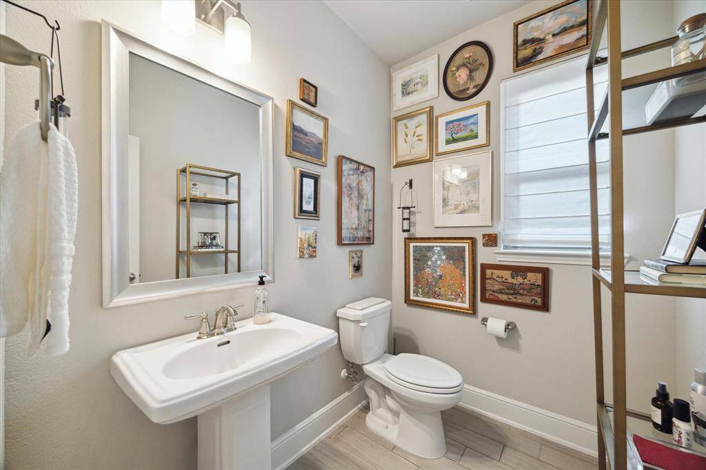 This bathroom features a sleek pedestal sink and modern toilet, complemented by a wall adorned with eclectic artwork. It has a large mirror, contemporary lighting, and a window with natural light.