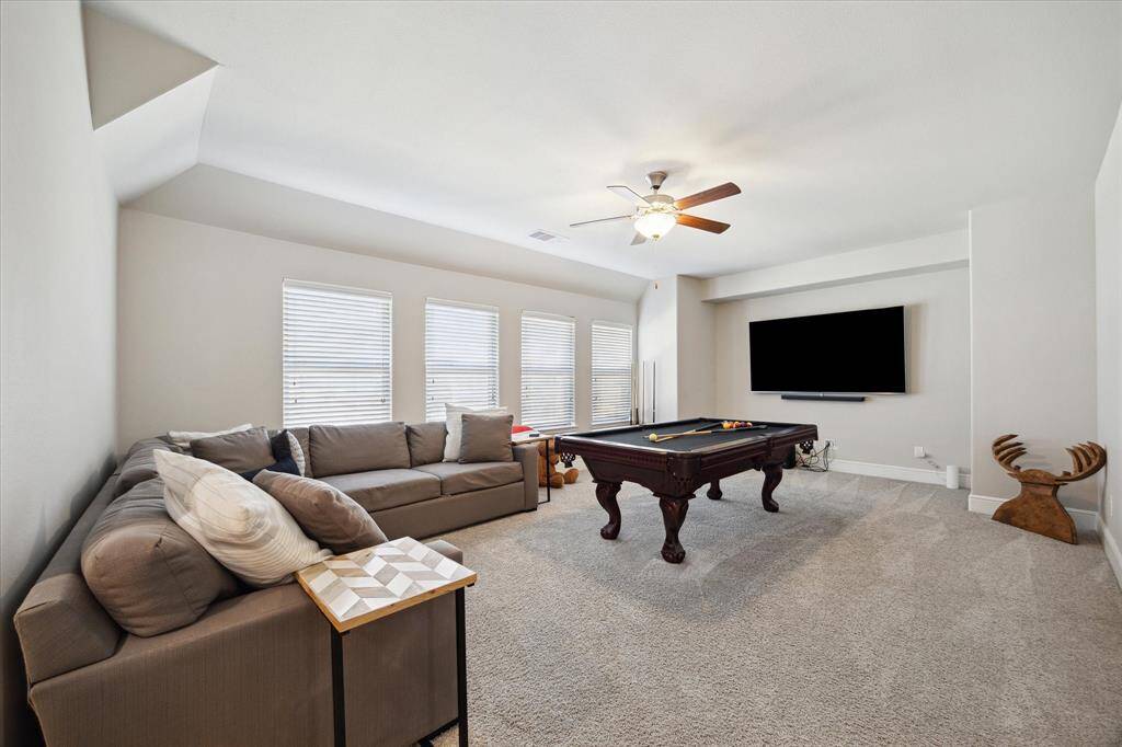 This spacious entertainment room features a plush sectional sofa, a pool table, and a wall-mounted TV, with natural light streaming in through multiple windows. The neutral tones and ceiling fan add comfort and style, making it an ideal space for relaxation and fun.
