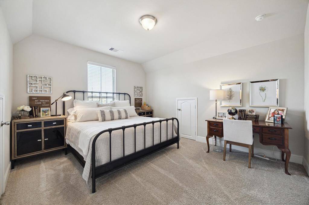This is a cozy, well-lit bedroom featuring a metal-framed bed, plush carpeting, and neutral walls. It includes a side table and a classic wooden desk with a chair, providing a comfortable and stylish space.