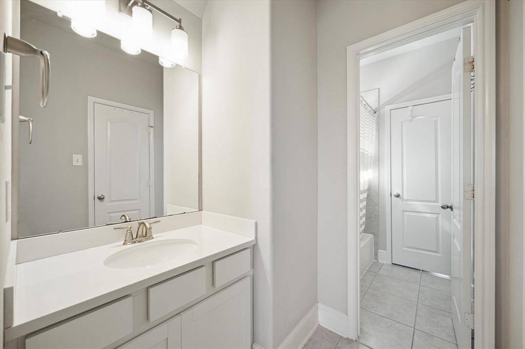 The Hollywood bathroom features a modern design with a large mirror above a white vanity, sleek fixtures, and ample lighting. A tiled floor leads to a door opening into a shower area, offering a clean and bright space.