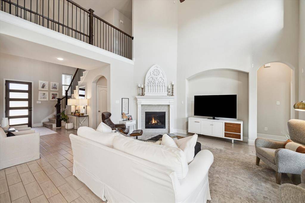 This spacious living room features high ceilings, a cozy fireplace, and abundant natural light. It includes a modern staircase leading to an upper level, elegant furnishings, and a wall-mounted TV. The open layout is perfect for entertaining and relaxation.