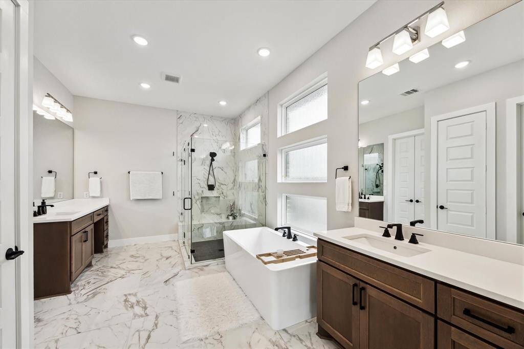This luxurious primary bathroom features a modern design with dual sinks, a spacious glass-enclosed shower, a standalone bathtub, and elegant flooring. Large mirrors and ample lighting create a bright atmosphere.