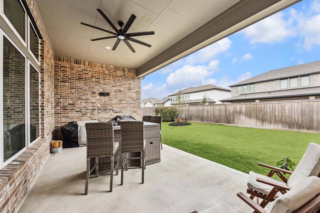 A spacious covered patio with a ceiling fan, brick wall, and outdoor seating. It overlooks a large, well-maintained backyard enclosed by a fence, perfect for relaxation and entertaining.