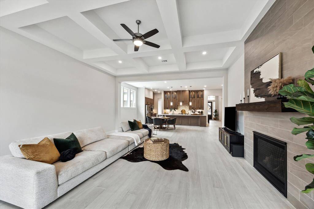 Modern open-concept living area with a cozy fireplace, elegant coffered ceiling, and ample natural light. The space flows seamlessly into a sleek kitchen with warm cabinetry and a large island, ideal for entertaining.
