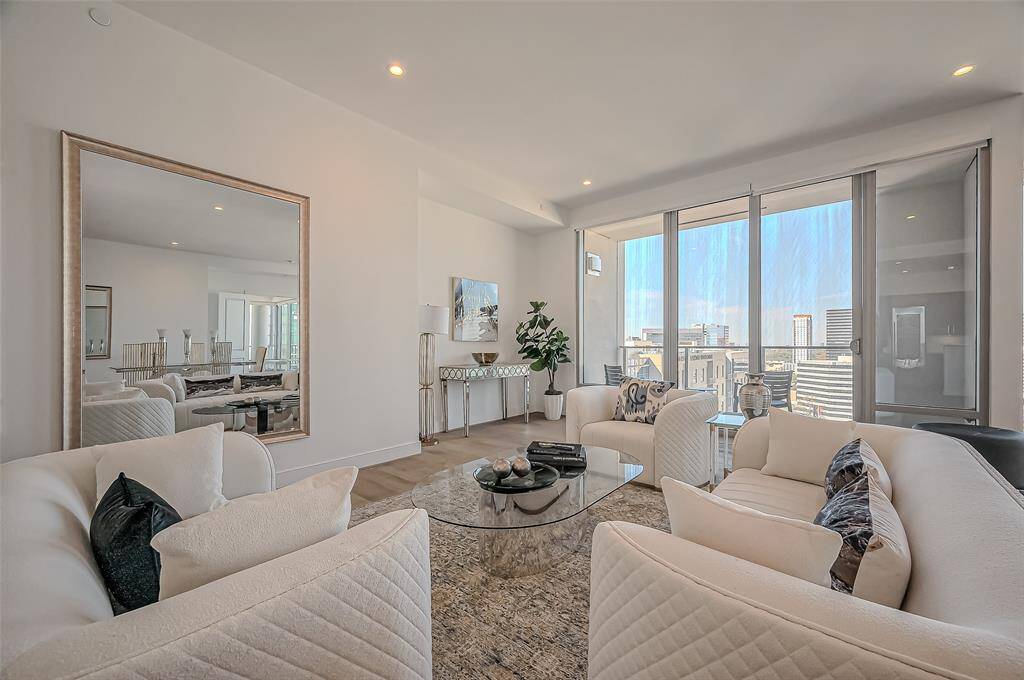 Bright and spacious living room with large windows offering city views. Features elegant white sofas, a glass coffee table, and contemporary decor. Perfect for entertaining or relaxing.