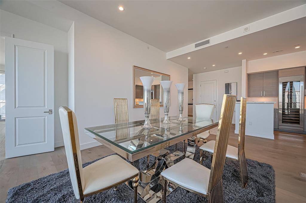 Modern dining area with a sleek glass table and stylish chairs on a plush rug. The open and bright space, with recessed lighting and a view into a contemporary kitchen featuring stainless steel appliances and dark cabinetry.