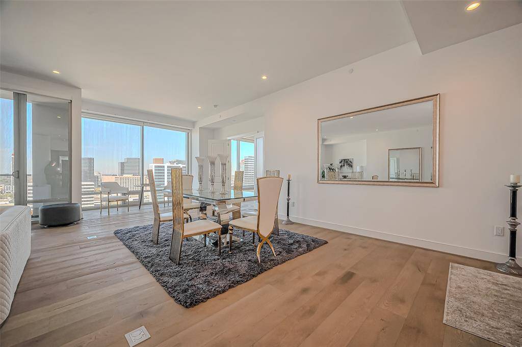 Dining area features a spacious, open layout with large windows offering city views. It has wooden flooring, a glass-top dining table, and a large wall mirror enhancing the room's brightness and openness.