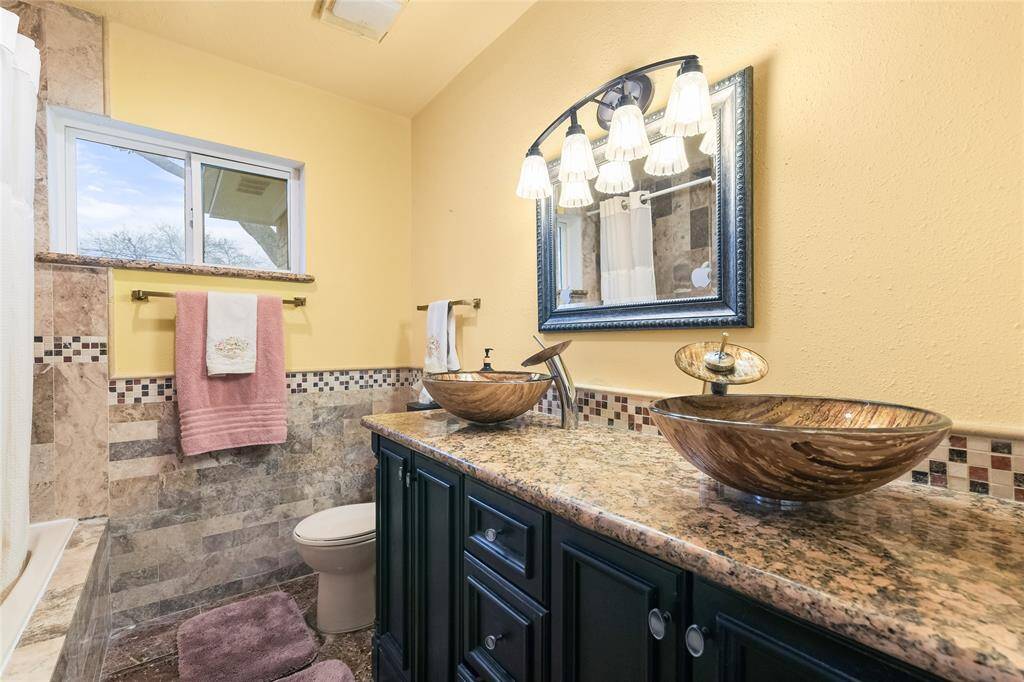 Step into this beautifully designed spa-like bathroom, where elegance meets functionality. The granite-topped double vanity with vessel sinks adds a touch of sophistication, complemented by sleek modern faucets and a framed mirror with stylish lighting. The rich tilework with mosaic accents creates a warm and inviting ambiance, while the contrasting dark cabinetry provides ample storage. A large window allows natural light to brighten the space, enhancing its cozy yet upscale feel.