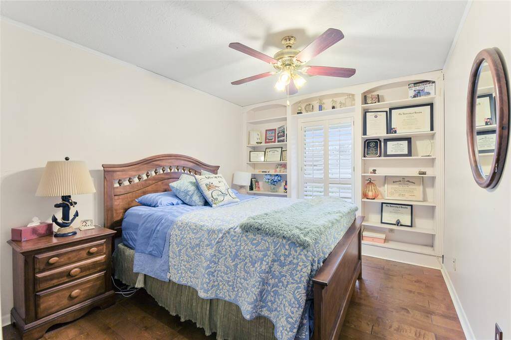 Featuring rich hardwood flooring, a stylish ceiling fan, and a neutral color palette, this space exudes warmth and relaxation. A standout feature is the custom built-in shelving, providing ample space for books, décor, or cherished keepsakes. The large window with plantation shutters allows for plenty of natural light while offering privacy.