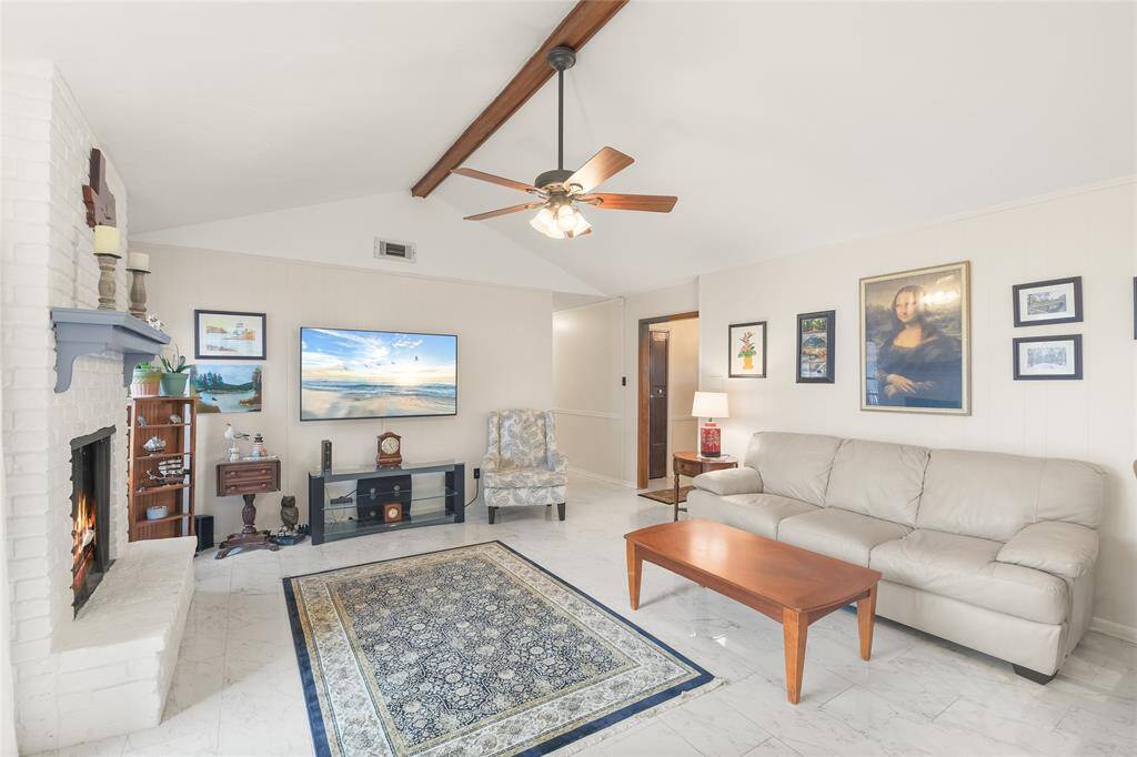 The vaulted ceiling with an exposed wood beam adds architectural charm, while the ceiling fan and recessed lighting ensure a cozy and well-lit ambiance. A white brick fireplace serves as a beautiful focal point, perfect for gathering around on cool evenings. The neutral color palette and elegant tile flooring create a modern and timeless aesthetic.