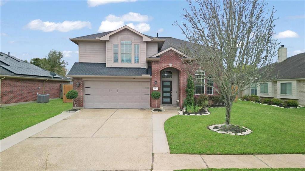 This photo shows a two-story brick home with a well-maintained front yard and driveway. The house features a two-car garage, arched entryway, and a prominent front window. The lawn is landscaped with a tree and shrubs, adding curb appeal.