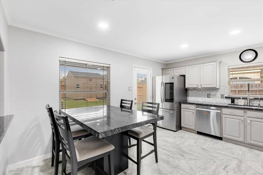 This kitchen boasts modern stainless steel appliances, sleek white cabinetry. Natural light floods in through two windows and a glass door, enhancing the bright and inviting atmosphere.