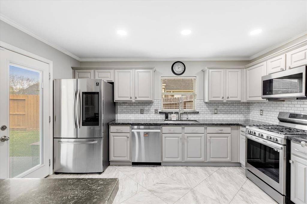 This kitchen features modern stainless steel appliances, light gray cabinetry, and a sleek subway tile backsplash. It has a spacious layout with ample counter space and a window above the sink for natural light. The flooring is a stylish, light-colored tile, and the room opens to the backyard through a glass door.