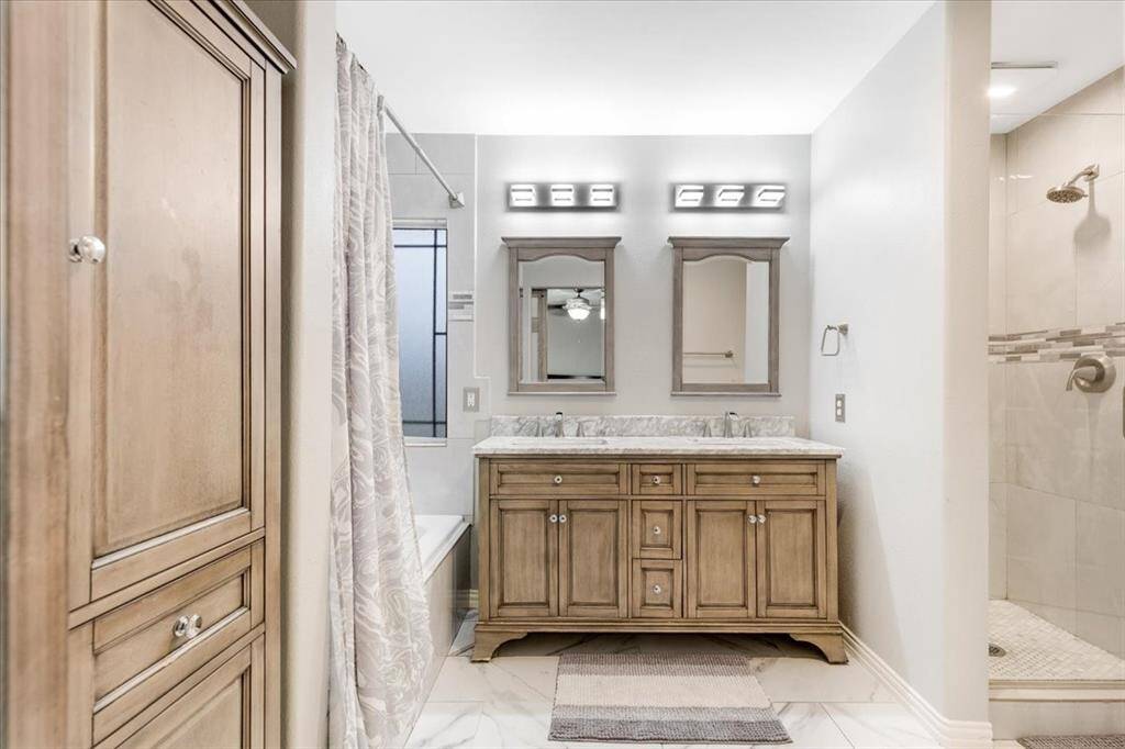 This bathroom features a dual-sink vanity with ample storage, elegant lighting, and a neutral color palette. It includes a separate shower with a glass door and a cozy tub, creating a comfortable and functional space.