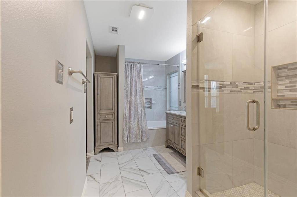 This modern bathroom features a spacious glass-enclosed shower with stylish tile accents, a separate bathtub with a patterned curtain, and a sleek vanity. The room is enhanced by elegant cabinetry and light-colored flooring, creating a bright and inviting atmosphere.
