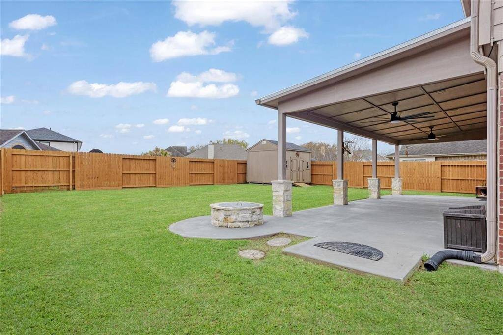 Spacious backyard featuring a covered patio with ceiling fans, perfect for outdoor gatherings. It includes a stone fire pit and is enclosed by a wooden privacy fence, offering a well-maintained lawn and a storage shed in the background.