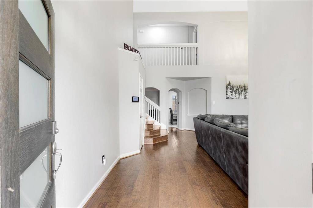 This photo showcases the entrance of a home with a modern wooden door, leading into an open living space with high ceilings. Natural light enhances the airy feel, and there's a balcony-style upper hallway visible.