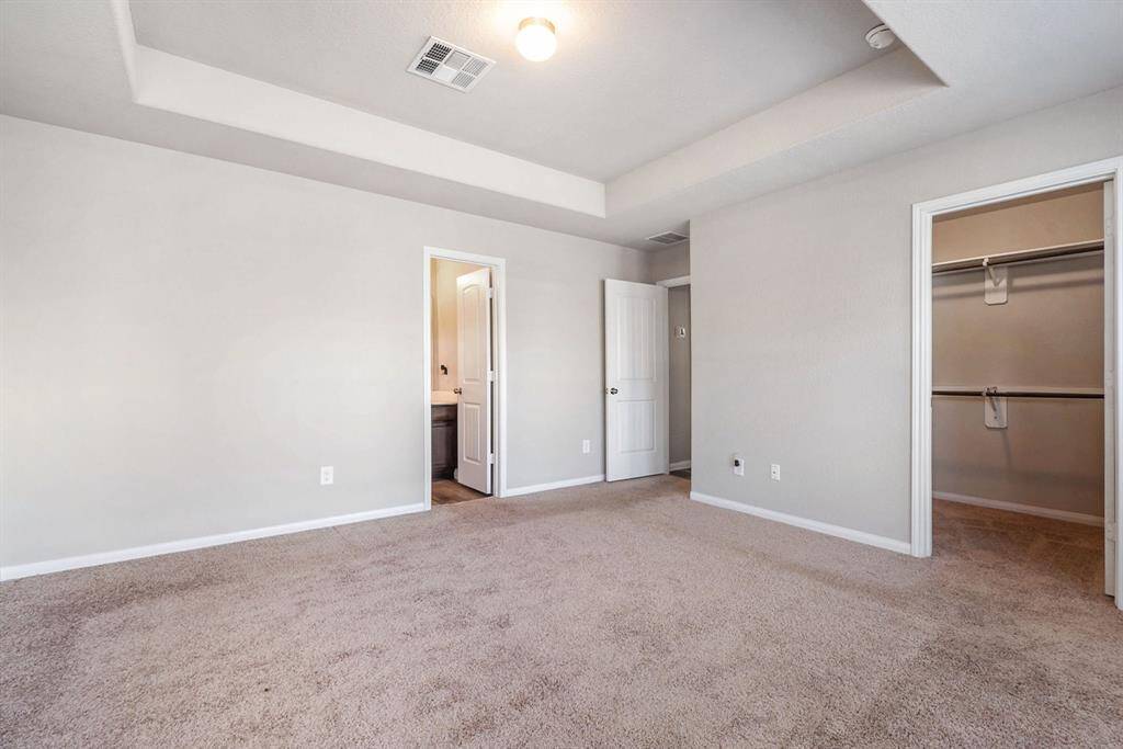 Spacious primary bedroom with neutral-toned carpet and walls. It features a tray ceiling with recessed lighting, a doorway leading to a bathroom, and a walk-in closet, offering ample storage space.